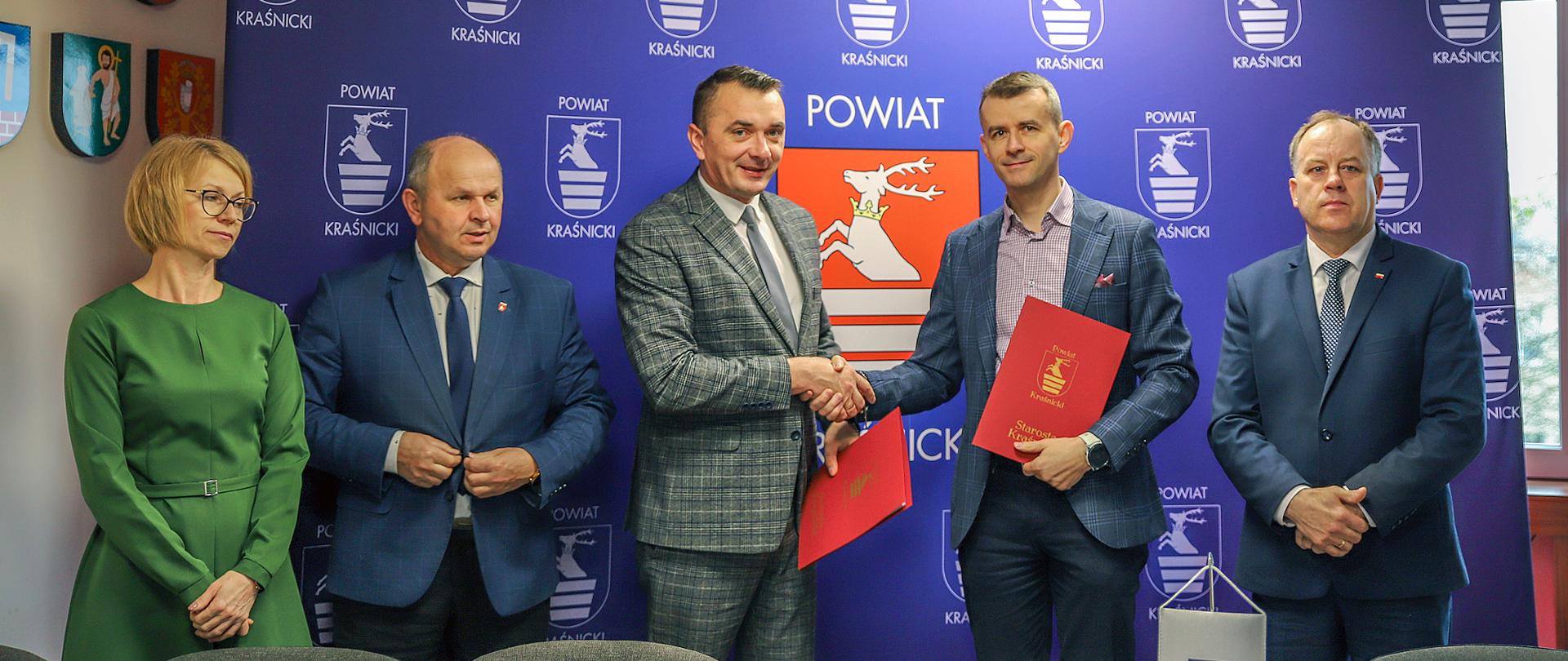 Zdjęcie przedstawia moment przekazania podpisanych egzemplarzy umowy. Na pierwszym planie znajduje się stół z krzesłami, za którym obok siebie stoi pięć osób: Skarbnik Powiatu Iwona Szymańska, Wicestarosta Kraśnicki Andrzej Misztal, Starosta Kraśnicki Paweł Kudrel, Wiceprezes Zarządu WOD-BUD Mirosław Gucwa, Radny Rady Powiatu Krzysztof Skórski. Starosta Kraśnicki oraz Wiceprezes Zarządu ściskają sobie dłonie przekazując podpisane umowy. W tle za zebranymi znajduje się niebieska ścianka promocyjna Powiatu Kraśnickiego z wizerunkami herbu.