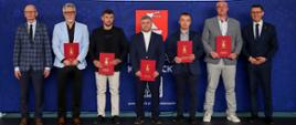 5 nagrodzonych trenerów stojących na tle granatowej ścianki reklamowej Starostwa Powiatowego w Kraśniku z herbem Powiatu Kraśnickiego pośrodku. Trenerzy trzymają w rękach bordowe teczki z herbem Powiatu Kraśnickiego i napisem Starosta Kraśnicki. Pierwszy od lewej strony stoi Starosta Kraśnicki Zbigniew Dżugaj, zaś jako ostatni Przewodniczący Rady Powiatu w Kraśniku Jacek Dubiel.