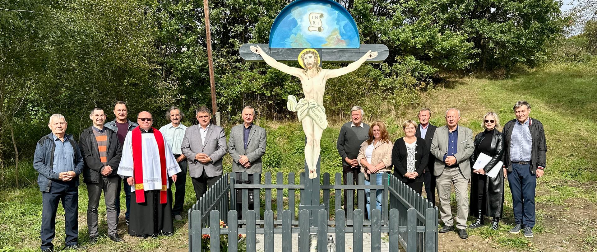 Na zdjęciu znajduje się grupa 15 osób, wśród których znajduje się Burmistrz Miasta, Ksiądz, Radni Miasta, a także lokalna społeczność. Przez grupą osób widoczny jest odnowiony krzyż.