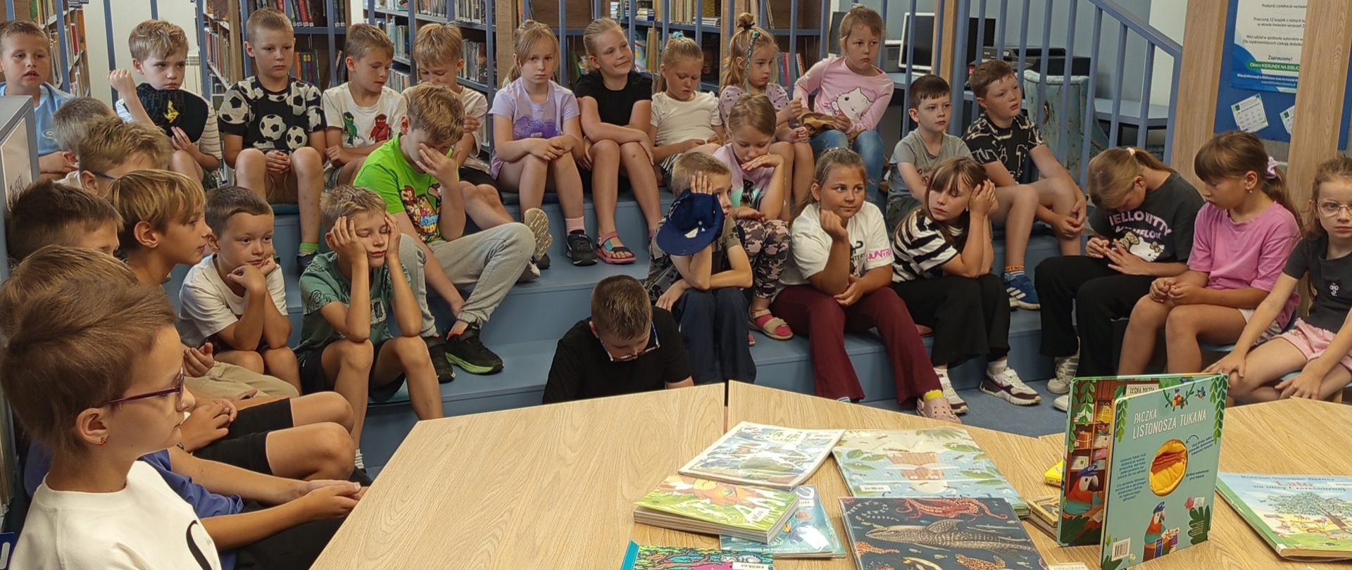 Dzieci podczas wakacyjnych zajęć w bibliotece siedzą na stopniowanych siedziskach i krzesłach wokół stołów, na których rozłożono kolorowe, ilustrowane książki. W tle znajdują się regały wypełnione książkami.