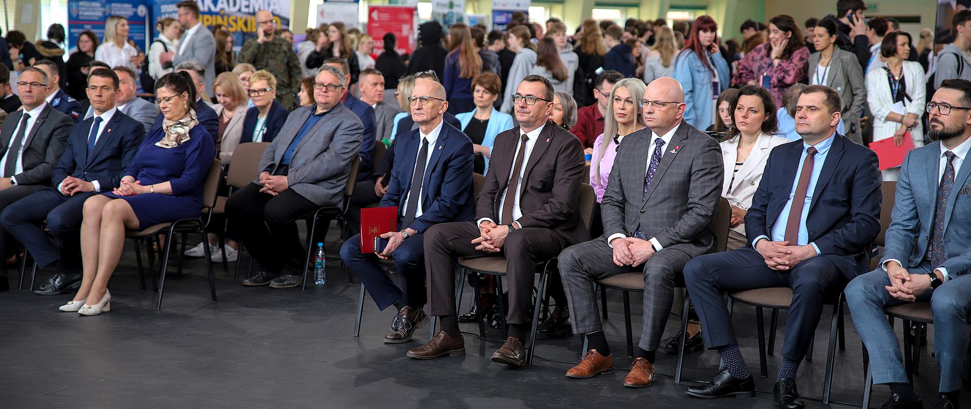 Zdjęcie przedstawia oficjalną część targów edukacyjnych, odbywających się w dużej hali sportowej. Pierwszy plan: W rzędzie na krzesłach siedzi grupa uroczyście ubranych osób – kobiet i mężczyzn w garniturach. To goście honorowi i organizatorzy. Siedzą na tle ciemnej podłogi, zwróceni w stronę, której nie widać w kadrze.
Drugi plan: Za siedzącymi osobami widać wypełnioną przestrzeń hali. Znajduje się tam tłum ludzi, głównie młodzieży, która przechadza się między stoiskami.
Tło: W głębi widoczne są kolorowe ścianki wystawiennicze, banery (m.in. z napisem „Akademia”) oraz elementy infrastruktury sportowej, jak kosz do koszykówki i wysoki, spadzisty sufit hali z oknami.