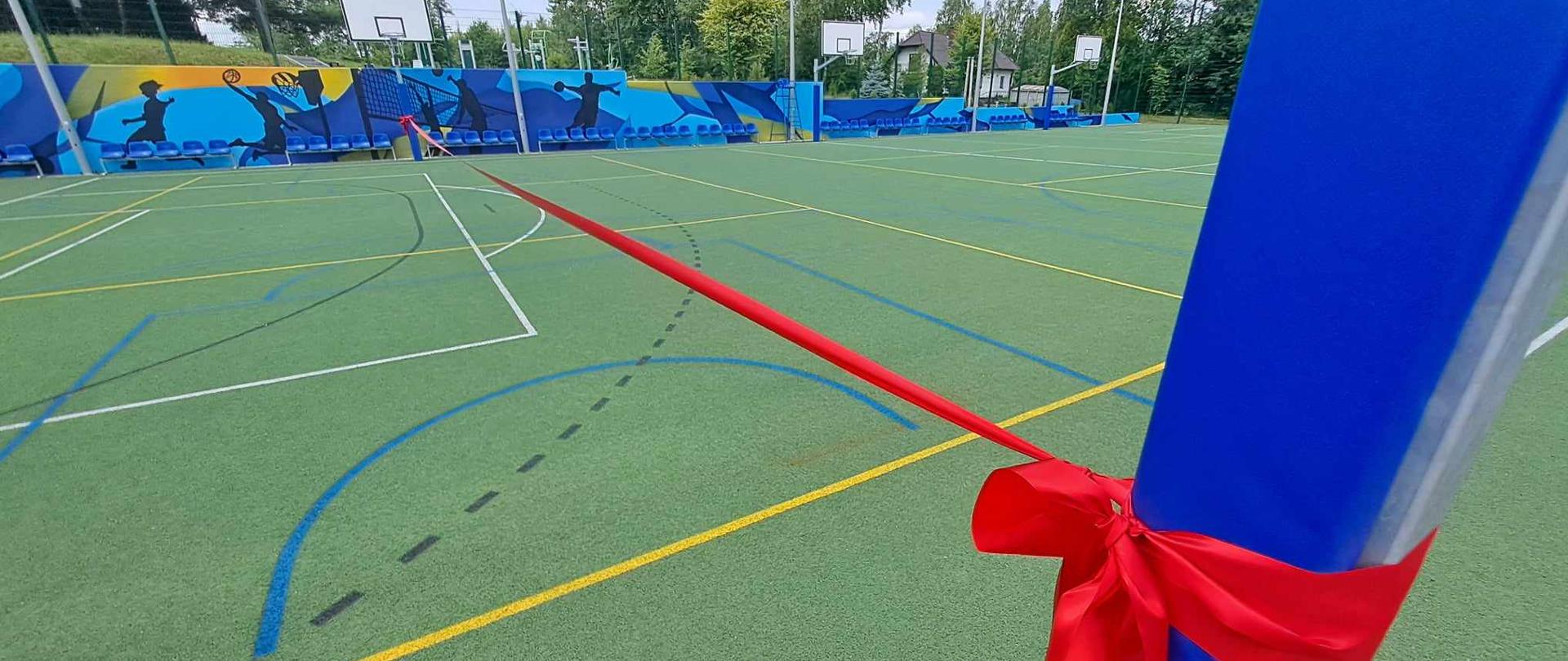 Na zdjęciu widzimy boisko sportowe z zieloną nawierzchnia poliuretanową przez środek boiska wisi czerwona wstęga. 