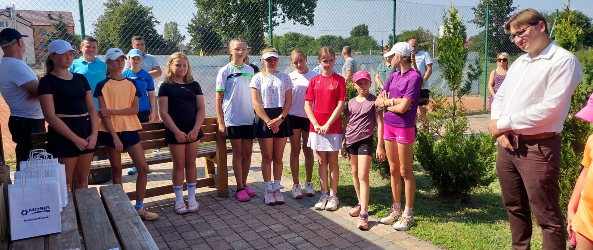 Uczestnicy turnieju stojący na tle kortu tenisowego. Pierwszy od lewej stoi Zastępca Dyrektora Miejskiego Ośrodka Sportu i Rekreacji w Kraśniku Łukasz Bielak