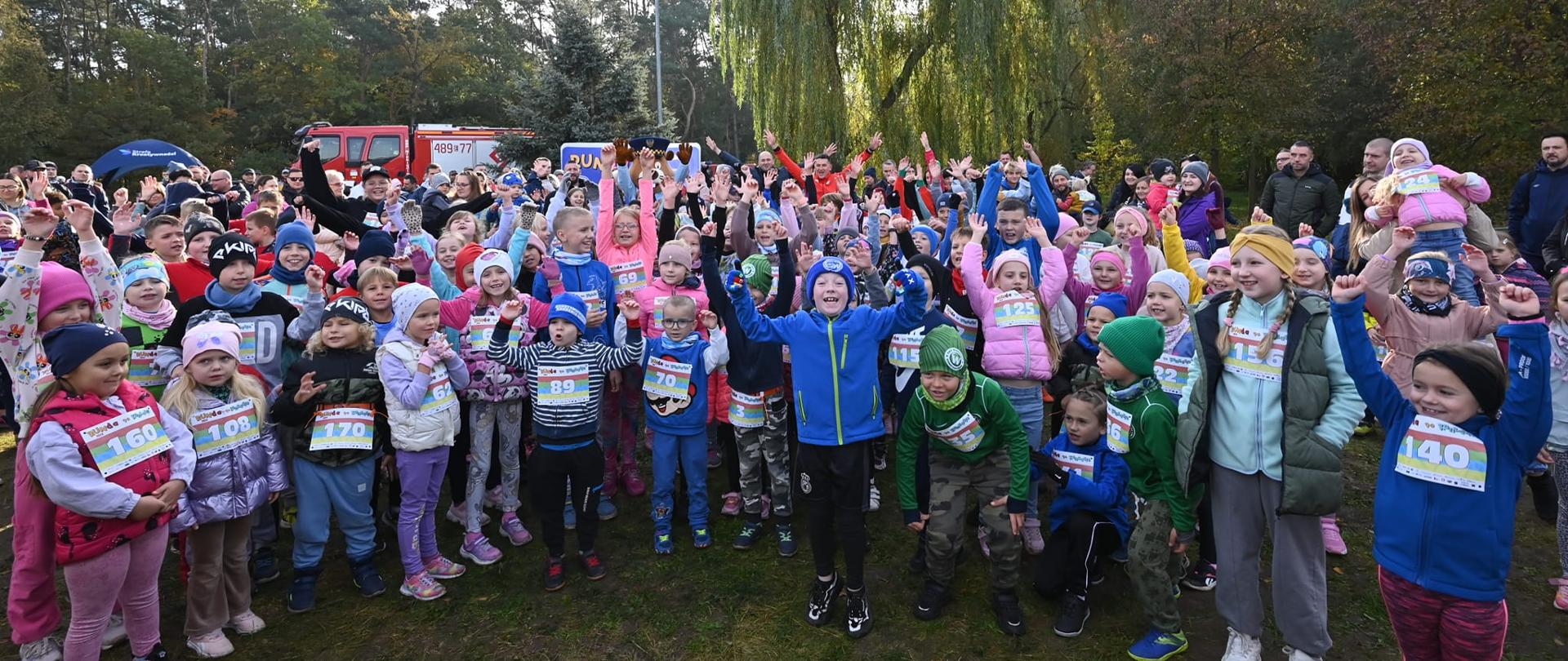 Grupa kilkudziesięciu dzieci w kolorowych strojach sportowych stoi na trawie, uśmiecha się i macha rękami do góry. Na piersiach mają przypięte numery startowe, co wskazuje na udział w biegu. W tle widoczni są opiekunowie oraz drzewa w jesiennej scenerii.