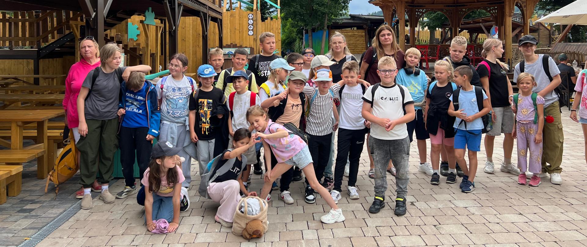Grupa dzieci wraz z opiekunami pozuje do wspólnego zdjęcia na terenie parku rekreacyjnego. W tle widoczne są drewniane konstrukcje, altana oraz otaczająca je zieleń.
