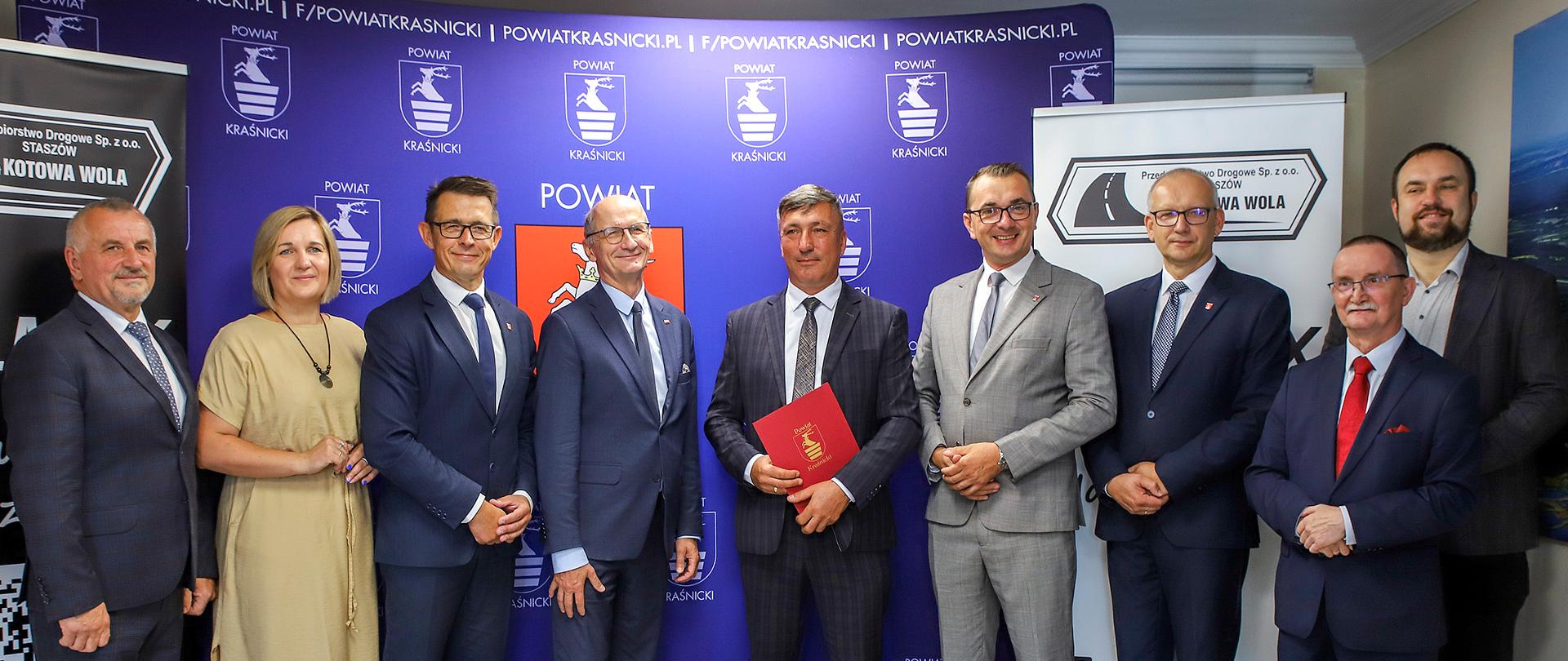 Pamiątkowe zdjęcie z okazji podpisania umowy. Na zdjęciu dziewięć osób stojących w rzędzie pozuje do zdjęcia na tle materiałów reklamowych powiatu kraśnickiego i przedsiębiorstwa budowlanego.