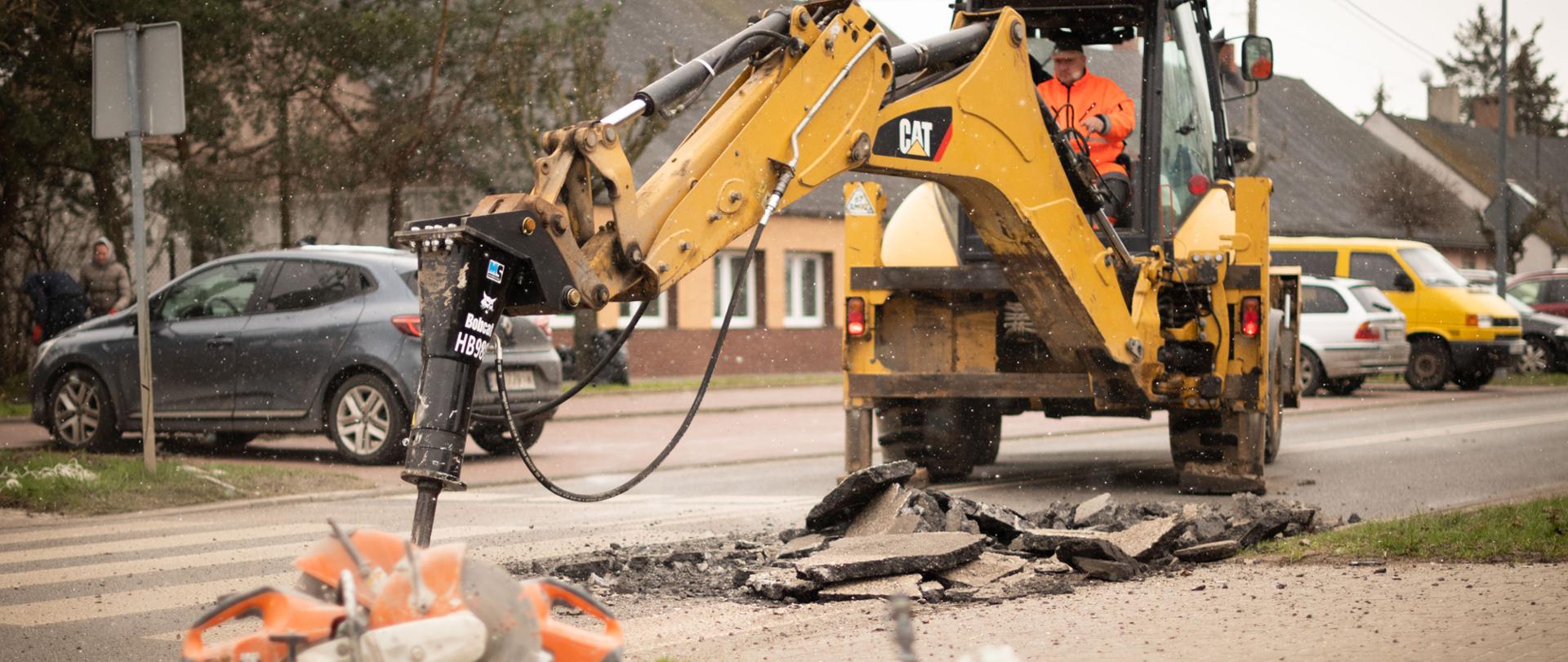 Zdjęcie przedstawia traktor, w którym siedzi mężczyzna w żółtym stroju. Trwają prace remontowe na ulicy, a na boku widać łopaty i inne narzędzia. W tle znajdują się zaparkowane samochody.