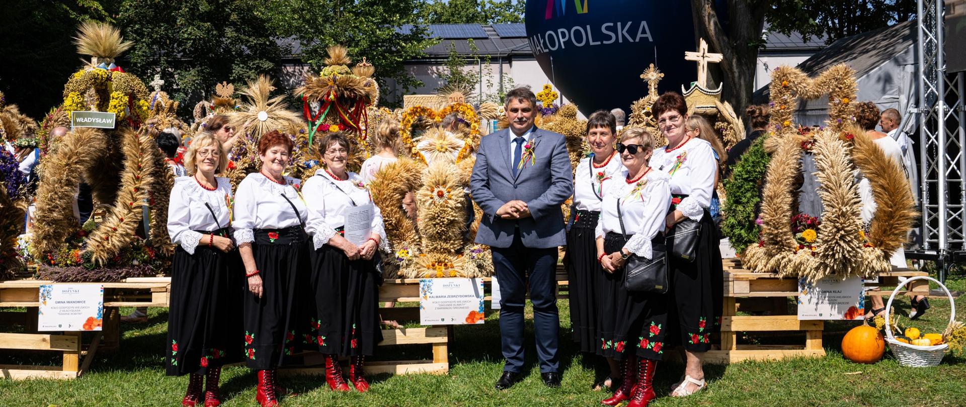 Burmistrz i członkinie koła gospodyń wiejskich pozują do zdjęcia przy wieńcach dożynkowych, za nimi granatowy balon z logo Małopolski.