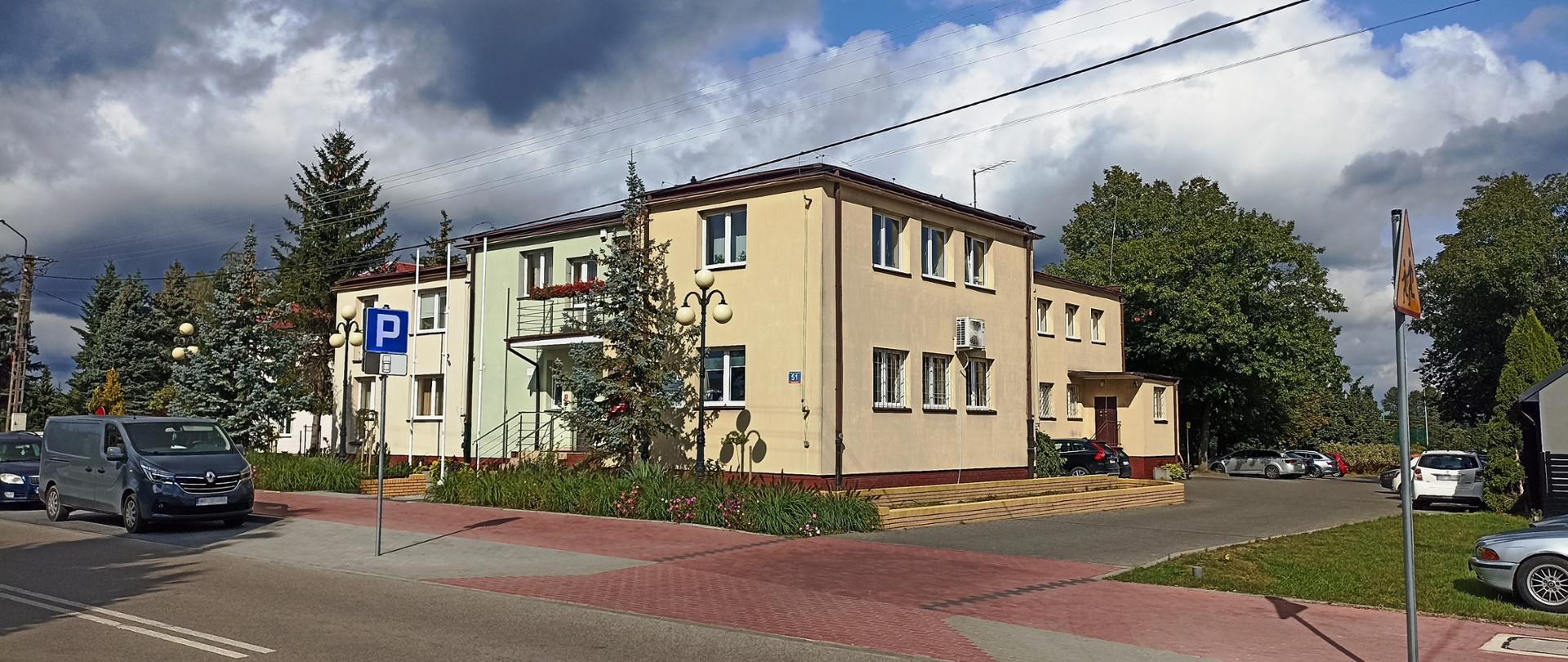 Jedno piętrowy budynek w kolorze ecru i jasnej zieleni. Przed nim rośnie drzewo iglaste.