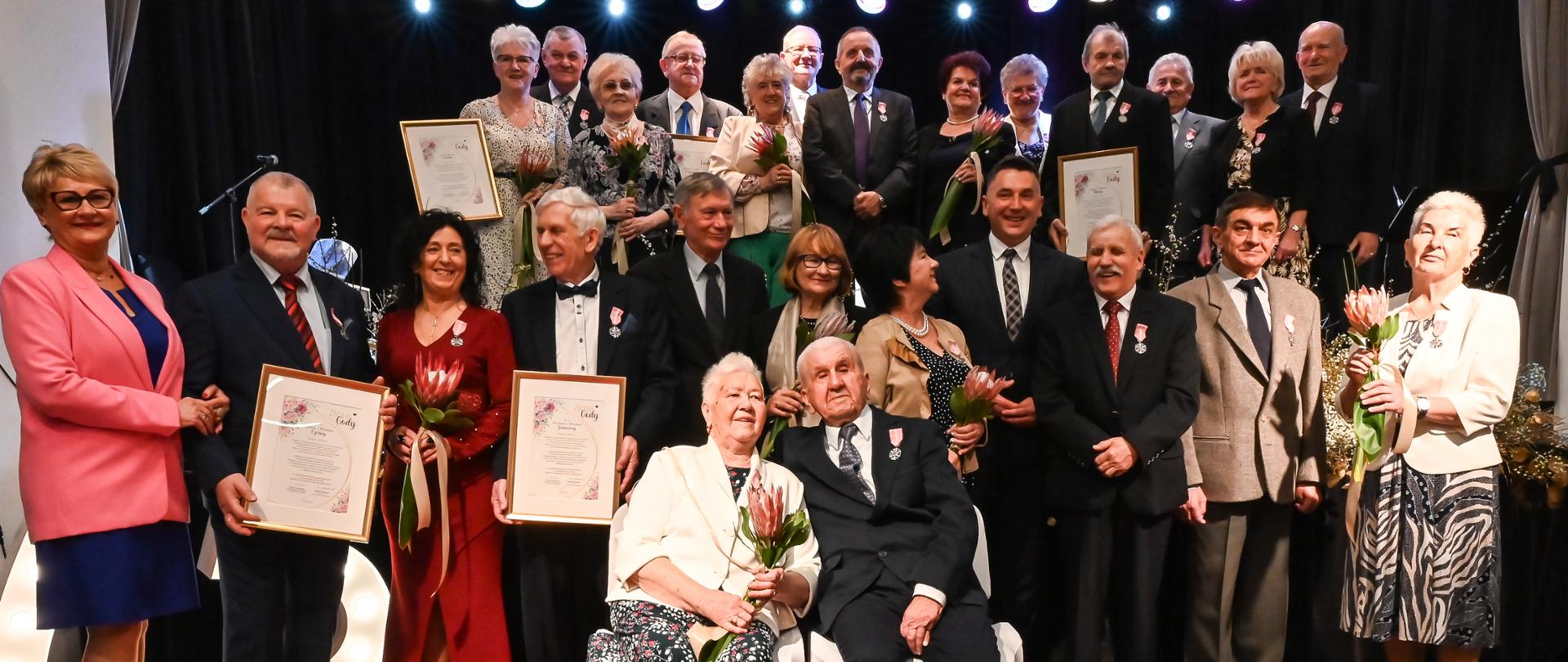 Zdjęcie grupowe odznaczonych medalami za długoletnie pożycie małżeńskie. Część par stoi na scenie, część pod sceną Miejskiego Ośrodka Kultury. Kilka osób trzyma w ręku dyplom. Z przodu najstarsza para siedzi na krzesłach.