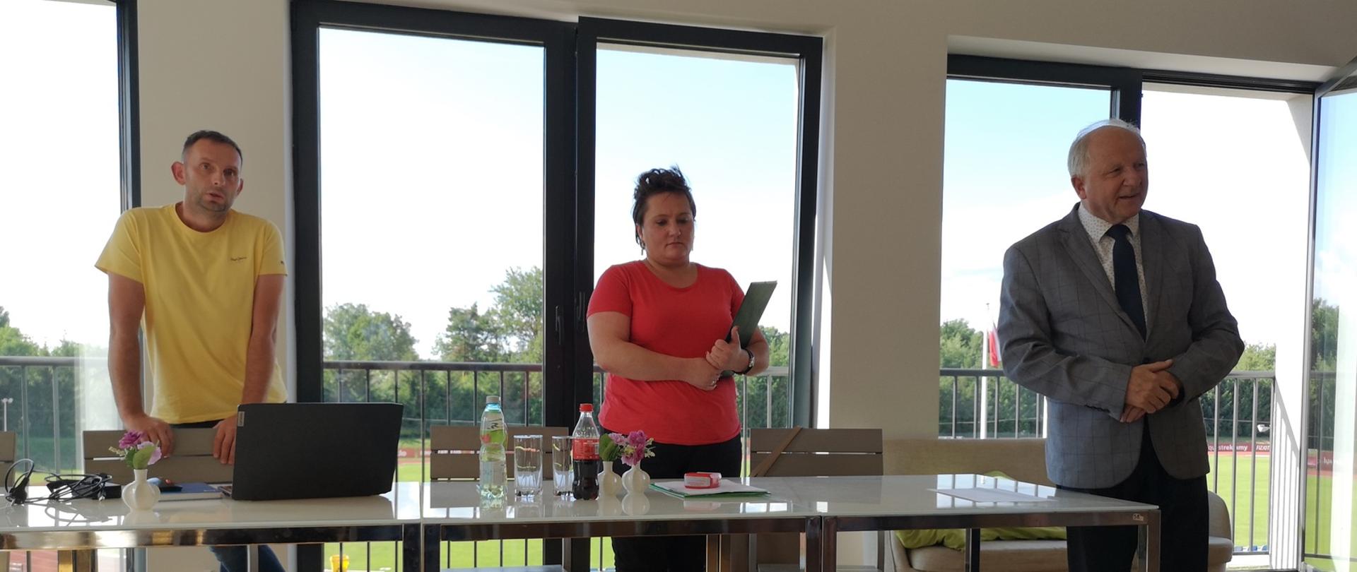 Trzy osoby prowadzące konferencję stoją przy stole, za nimi ściana z oknami tarasowymi