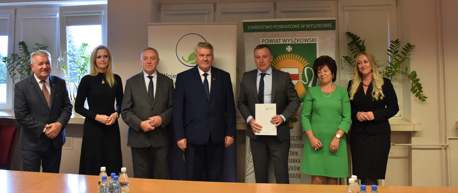 Dziewięć osób stoi przodem. Mężczyzna w środku trzyma w obu rękach plik dokumentów