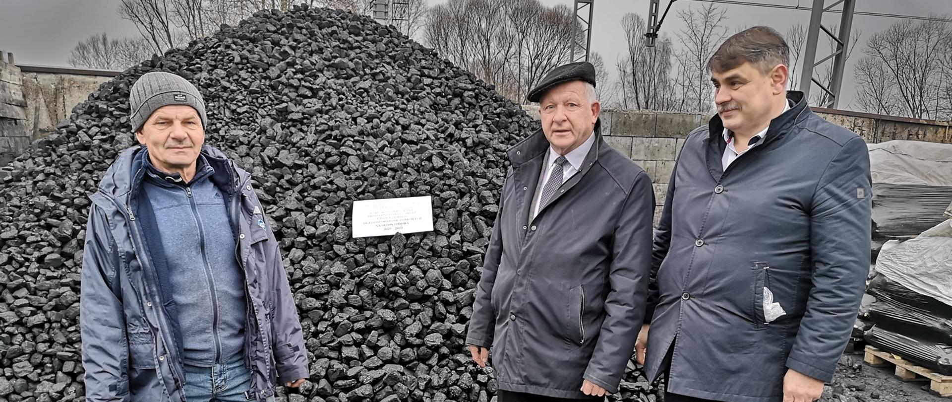 Burmistrz, Kierownik Referatu Gospodarki Komunalnej i właściciel składu stoją na placu przed pryzmą węgla. Na węglu kartka z informacją, że węgiel został zakupiony z programu rządowego