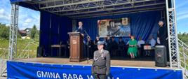 Na zewnętrznej scenie podczas obchodów 70-lecia OSP Bukowina-Osiedle mężczyzna przemawia z mównicy. Za nim siedzą strażacy w galowych mundurach oraz zaproszeni goście. Przed sceną stoi strażak w mundurze wyjściowym. Na froncie sceny widoczny jest napis „Gmina Raba Wyżna”, a w tle baner informujący o jubileuszu jednostki. Uroczystość odbywa się w słoneczny dzień.