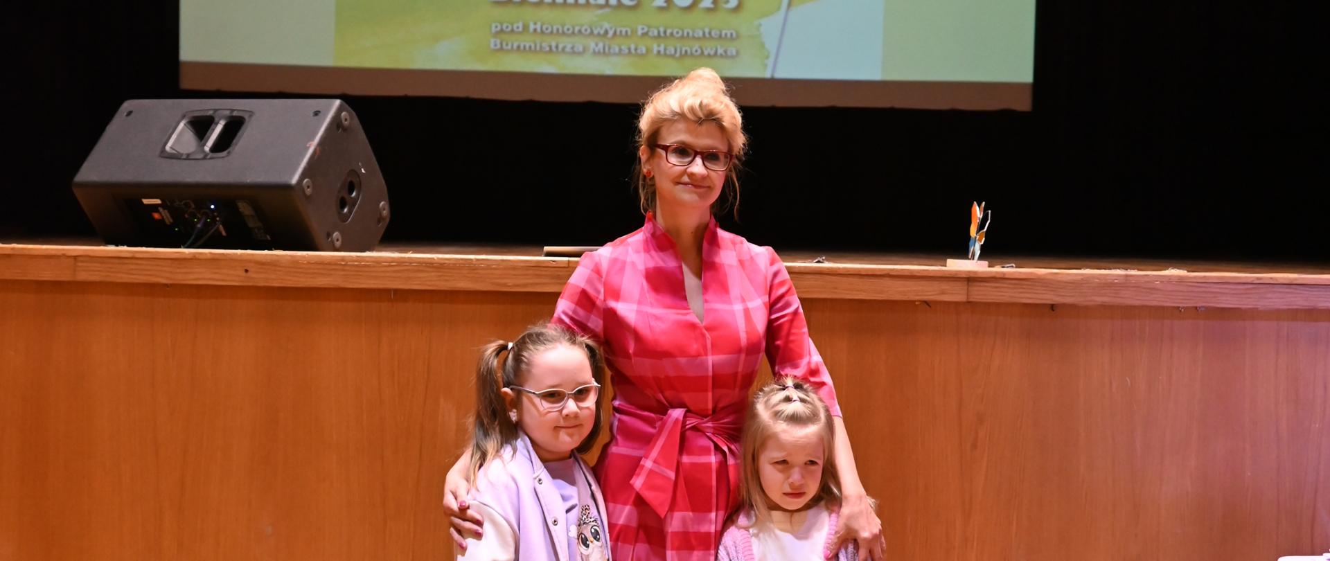 Dwie dziewczynki i nauczycielka pozują do zdjęcia. W tle rzutnik ekranu z plakatem wydarzenia