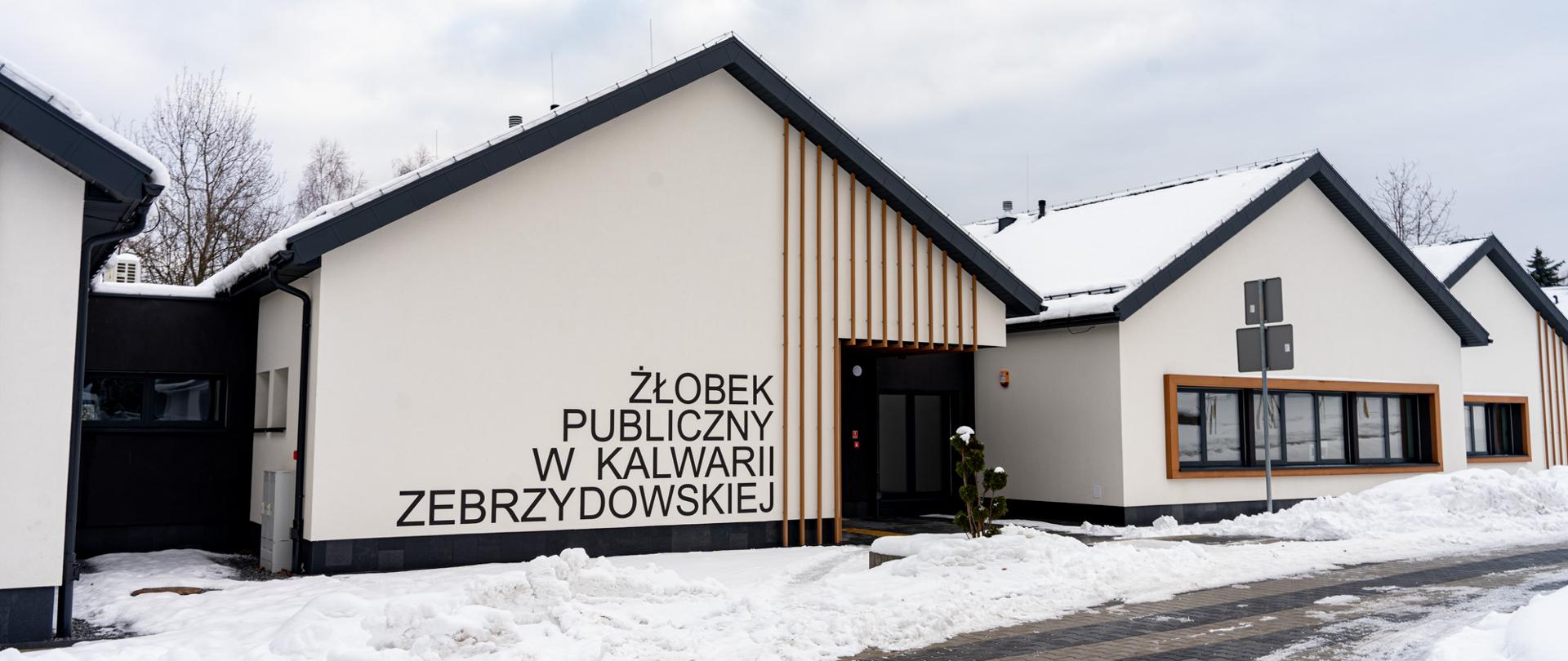 Nowoczesny budynek Żłobka Publicznego w Kalwarii Zebrzydowskiej zimą – jasna elewacja z napisem „Żłobek Publiczny w Kalwarii Zebrzydowskiej”, skośny dach pokryty śniegiem, przed wejściem odśnieżony chodnik i fragment drogi, w tle zimowe niebo.
