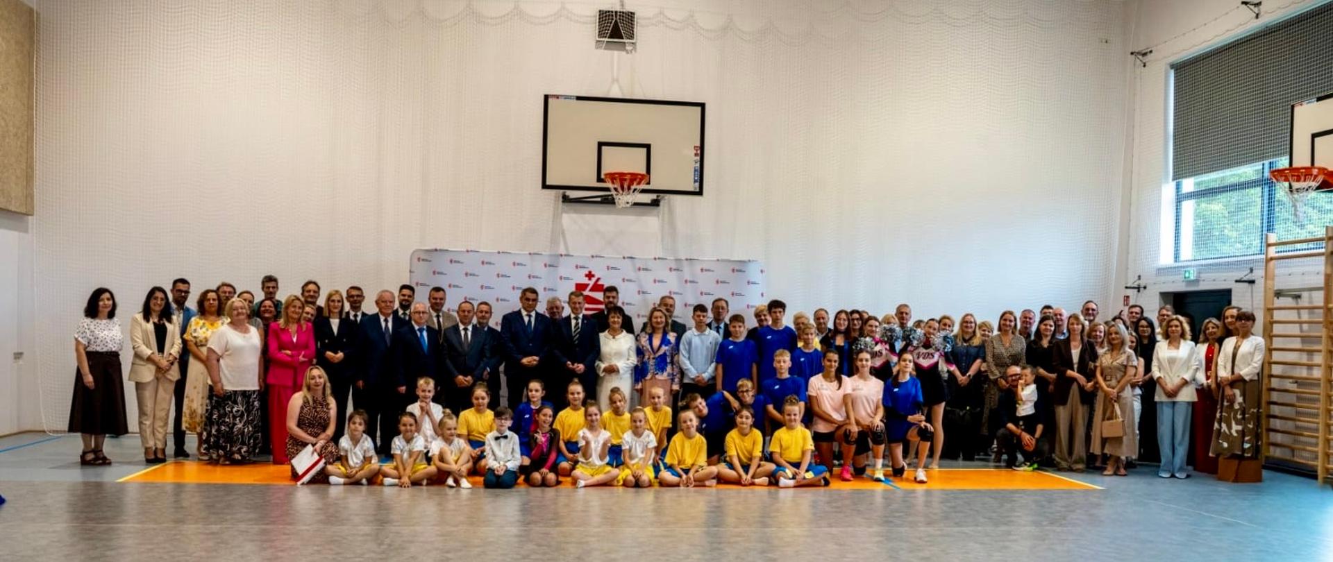 Zdjęcie grupowe uczestników uroczystego otwarcia nowej sali gimnastycznej w Zarzycach Wielkich. Na tle kosza do koszykówki i ścianki promocyjnej stoją zaproszeni goście, nauczyciele, rodzice i mieszkańcy, a przed nimi znajdują się uczniowie w strojach sportowych.