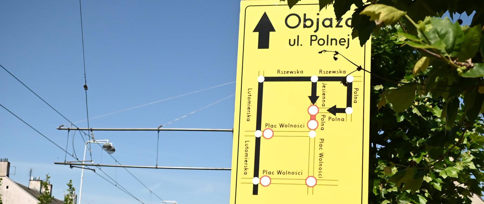 Żółta tablica informacyjna ustawiona przy ulicy przedstawia schemat objazdu w związku z zamknięciem fragmentu ul. Polnej. Na mapie zaznaczono trasę prowadzącą ulicami Rszewską, Lutomierską, Placem Wolności i Jesienną. W tle widoczna jest ulica z siecią trakcji tramwajowej, zabudową miejską oraz znakami drogowymi informującymi o prowadzonych pracach.