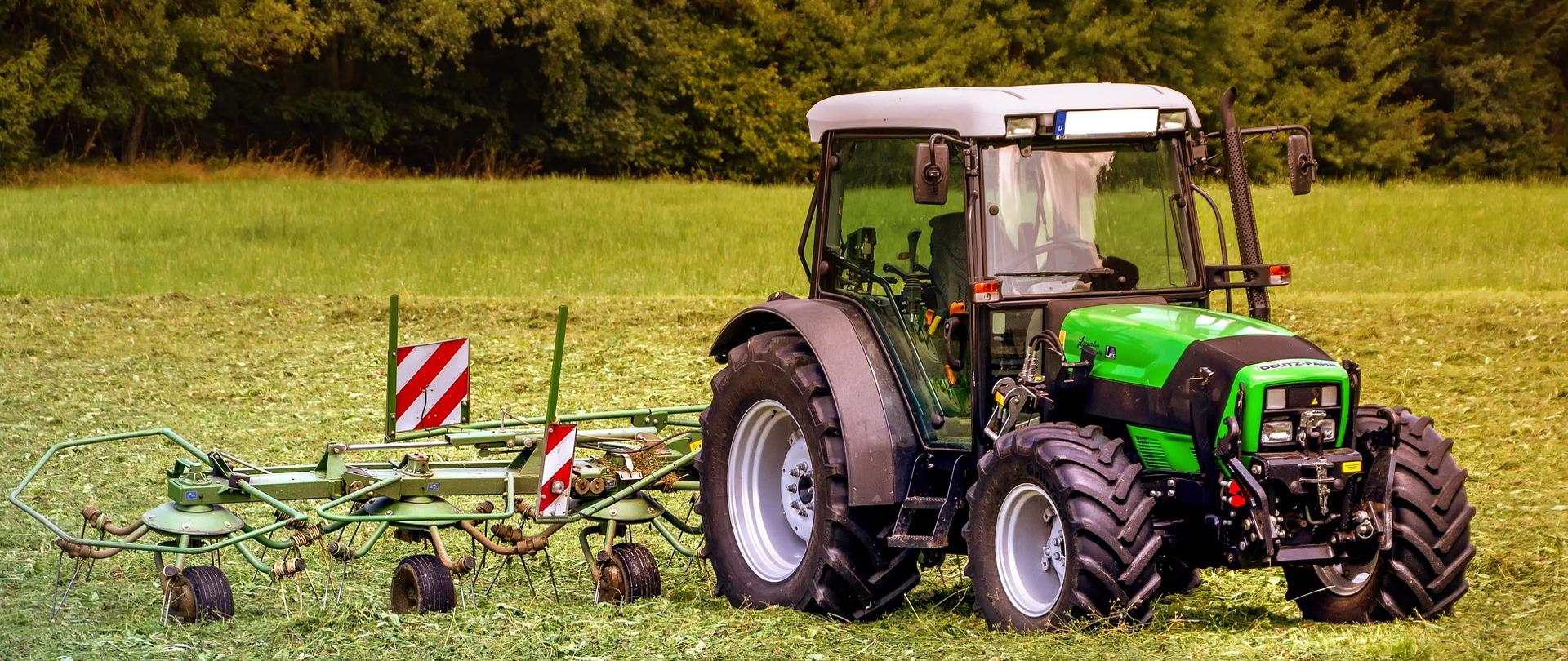 Traktor z podłączoną z tyłu kosiarką, stojący na wykoszonej łące, w tle gęsty las