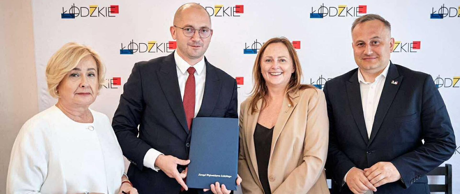 Cztery osoby stojące na tle ścianki z logotypem 'Łódzkie' i napisem 'Urząd Marszałkowski Województwa Łódzkiego'. Mężczyzna w garniturze i czerwonym krawacie trzyma granatową teczkę z napisem 'Zarząd Województwa Łódzkiego'. Wszyscy uśmiechnięci, w stroju formalnym
