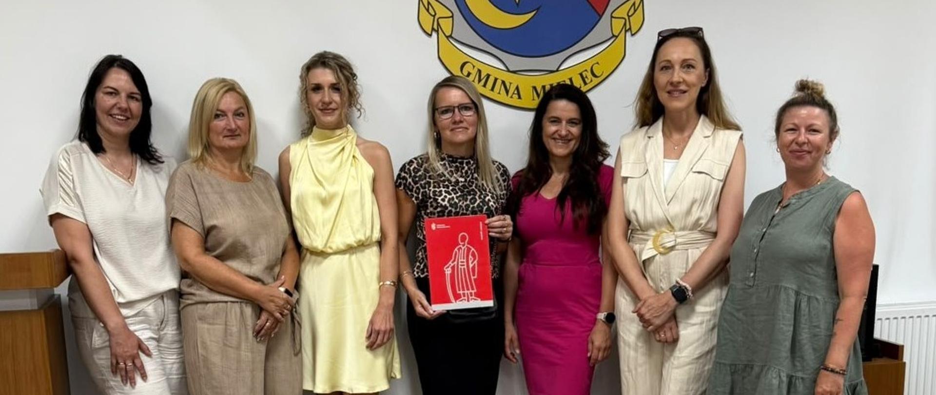 Na zdjęciu widać siedem kobiet stojących obok siebie w sali urzędu. W tle znajduje się duży herb Gminy Mielec umieszczony na białej ścianie. Jedna z kobiet trzyma czerwoną teczkę z białą grafiką przedstawiającą postać. Uczestniczki spotkania ubrane są w eleganckie, letnie stroje w jasnych i stonowanych kolorach. W pomieszczeniu widoczne są elementy wyposażenia sali, w tym drewniane meble oraz kamera monitoringu zamontowana pod sufitem. Zdjęcie ma charakter pamiątkowy i dokumentuje wspólne spotkanie lub wizytę studyjną.