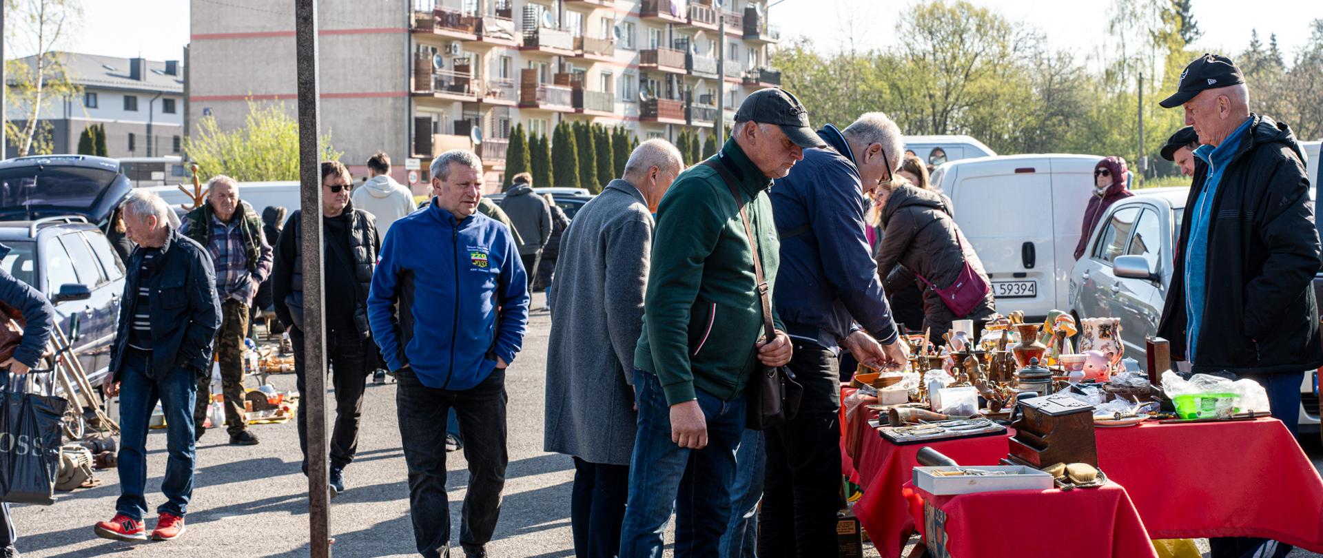 Grupa mieszkańców przegląda stoiska z przedmiotami z drugiej ręki podczas targu staroci na placu targowym. Na pierwszym planie stoły przykryte czerwonymi obrusami, na których wystawiono m.in. porcelanę, zegary, figurki i drobne antyki. W tle widoczne zaparkowane samochody oraz zabudowa mieszkaniowa. Nad częścią stoisk rozstawiony jest niebieski namiot, a cała scena odbywa się w słoneczny dzień.