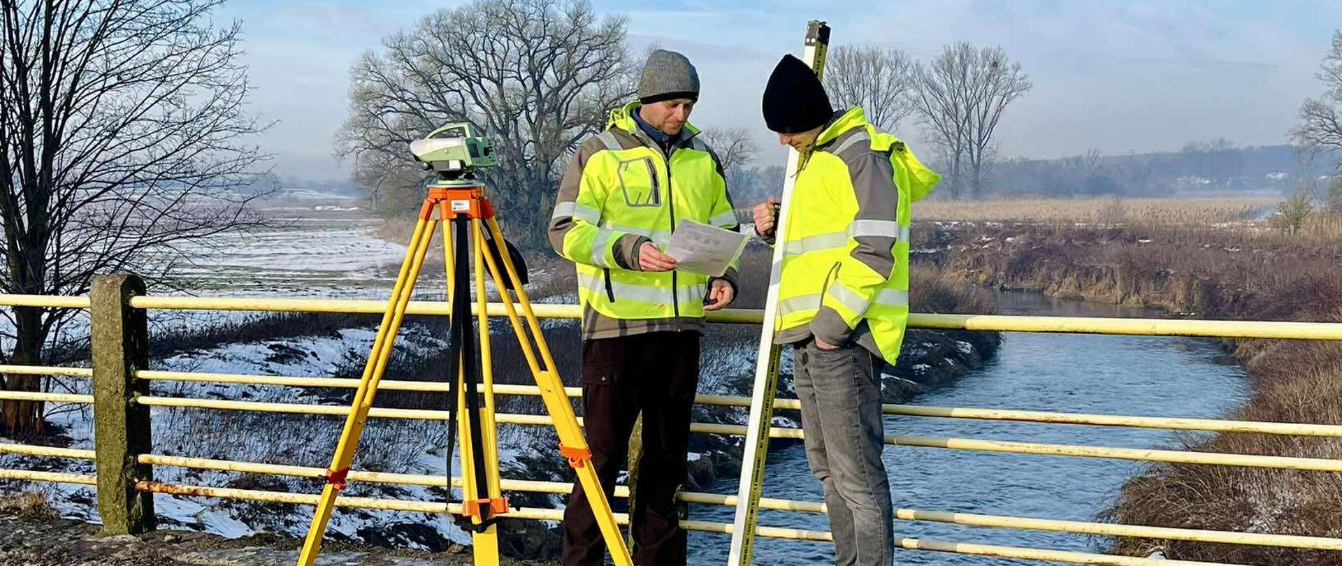 Dwóch geodetów w odblaskowych kurtkach stoi na moście nad rzeką i analizuje dokumentację pomiarową. Obok nich ustawiony jest statyw z urządzeniem geodezyjnym. W tle widoczna jest rzeka, zimowy krajobraz pól i drzew oraz błękitne niebo z lekkimi chmurami.