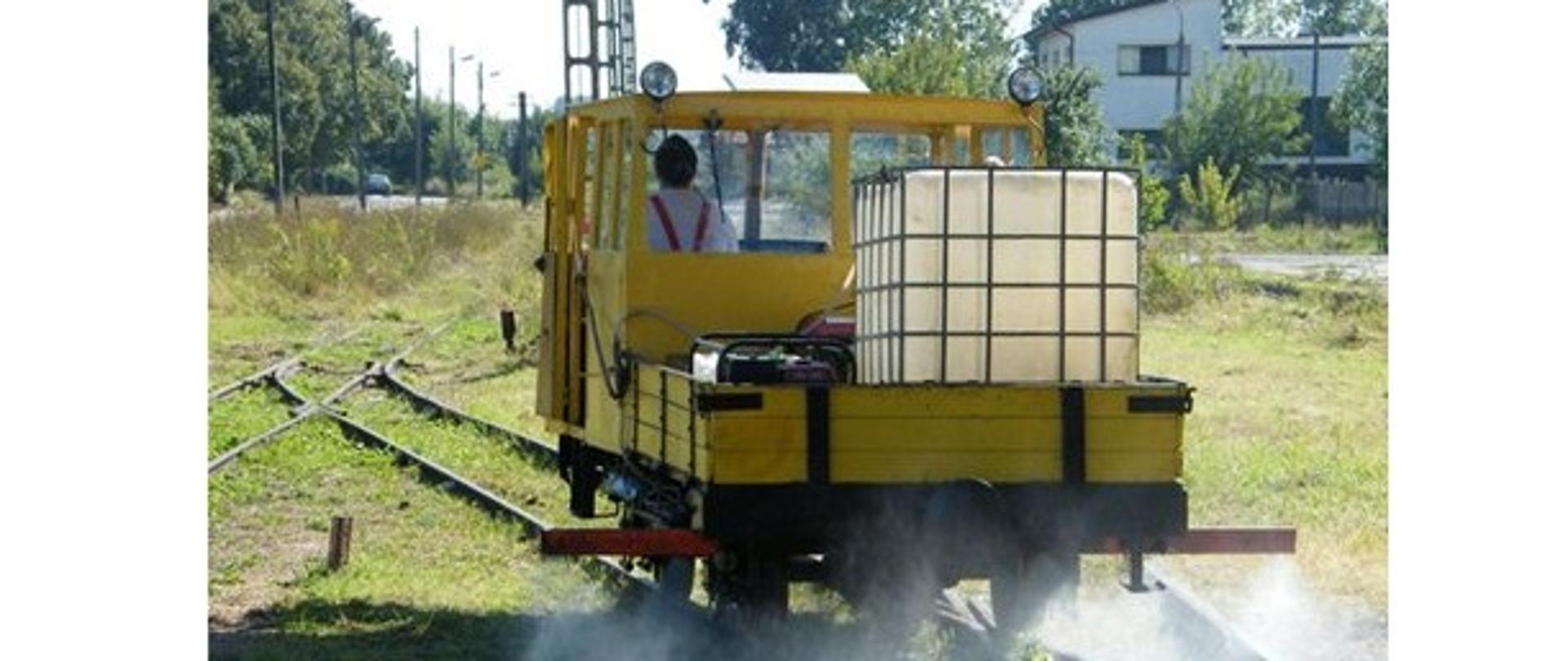 Wagon kolejowy podczas oprysku chwastobójczego. Wagon porusza się po szynach kolejowych z tyłu wagonu widać baniak ze środkiem chwastobójczym. Dookoła torów widać zieloną trawę i słupy energetyczne w tle po prawej stronie widać biały dom. 