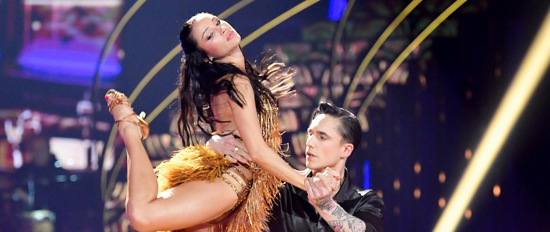 Para taneczna wykonuje dynamiczną figurę podczas występu w programie telewizyjnym. Tancerka w złotej, frędzlowanej sukni stoi na krześle i unosi nogę w powietrze, a partner w czarnym stroju podtrzymuje ją w trakcie układu.