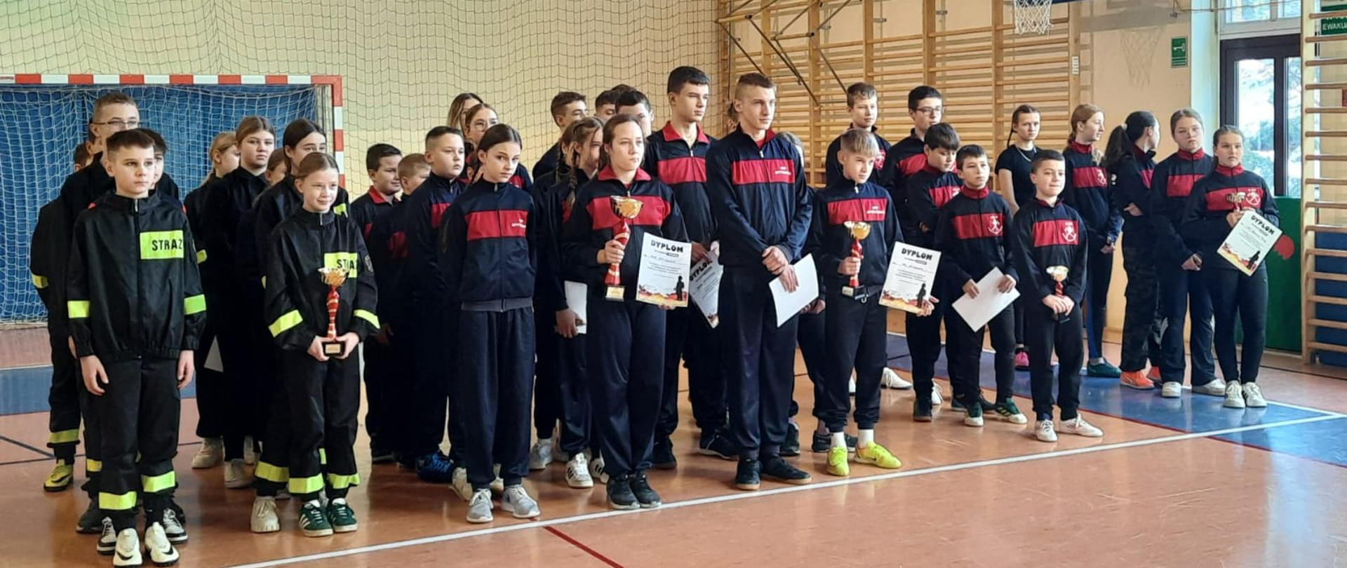 Grupa osób, drużyny młodzieżowe na sali gimnastycznej. Wszyscy ubrani w sportowe dresy. 