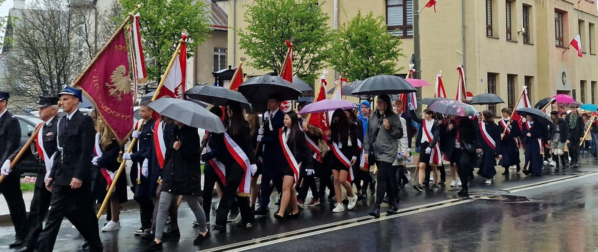 Prawym pasem mokrej od deszczu drogi maszeruje kondukt pocztów sztandarowych z flagami i parasolami. Za nimi budynek urzędu miasta ozdobiony biało-czerwonymi flagami.
