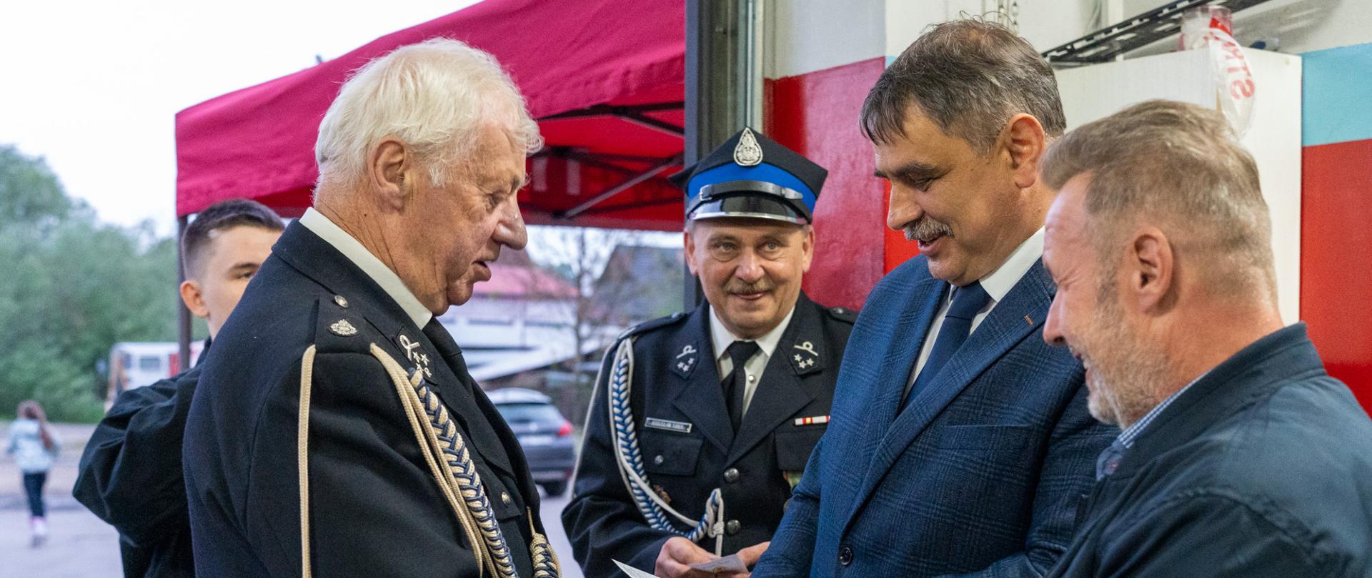 Burmistrz Miasta Kalwarii Zebrzydowskiej Tadeusz Stela podczas uroczystości Dnia Strażaka w OSP Brody wręcza gratulacje i rozmawia z druhami strażakami. W tle widoczni funkcjonariusze OSP w galowych mundurach oraz wnętrze remizy strażackiej. Fotografia wykonana podczas oficjalnej części wydarzenia.