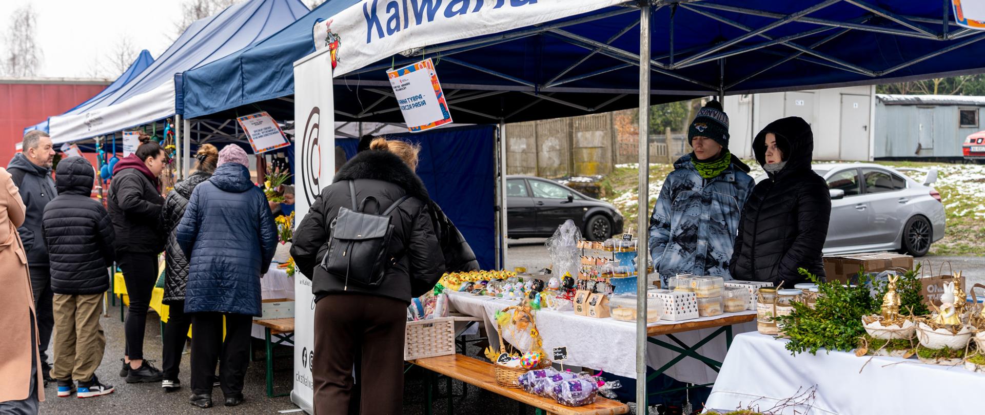 Rząd stoisk targowych pod niebieskimi namiotami z napisem „Kalwaria Zebrzydowska”, przy których mieszkańcy oglądają i kupują wielkanocne dekoracje, rękodzieło oraz słodkości; sprzedawcy stoją za stołami, a całość odbywa się na świeżym powietrzu w chłodny dzień.