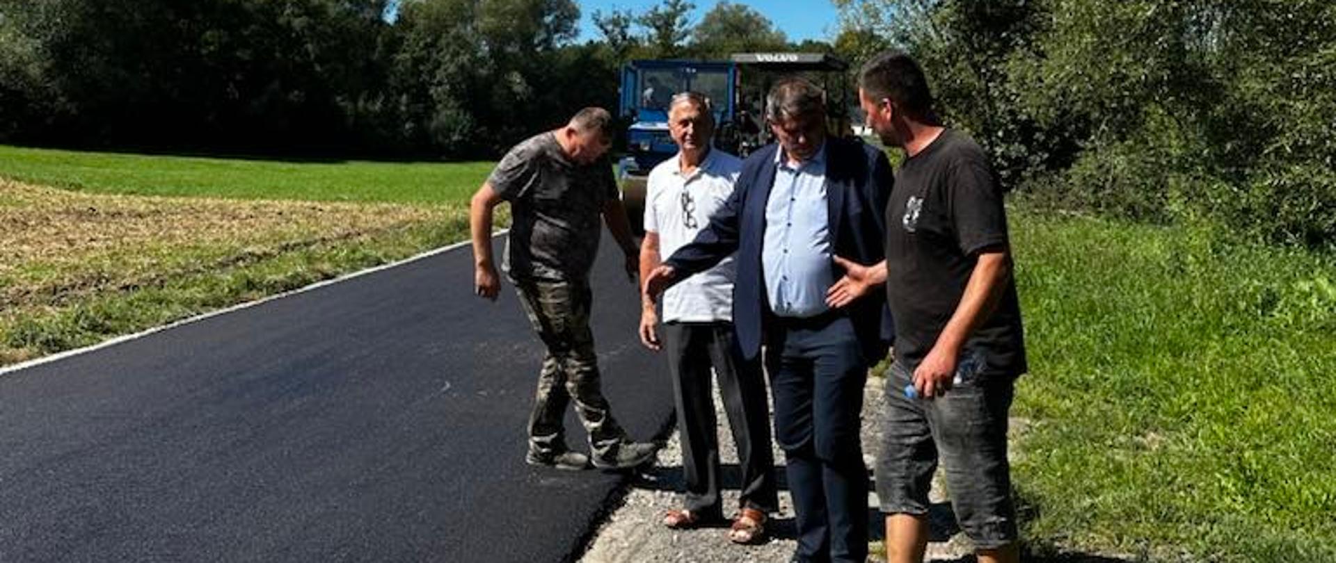 Na zdjęciu widać czterech mężczyzn stojących na nowo wyasfaltowanej drodze w słoneczny dzień. Dwóch z nich ubranych jest w ubrania robocze w kolorze moro, jeden ma jasną koszulę i ciemne spodnie, a jeden – marynarkę i koszulę. W tle znajdują się zielone pola, drzewa oraz fragment niebieskiego ciągnika. Niebo jest bezchmurne i intensywnie błękitne. 