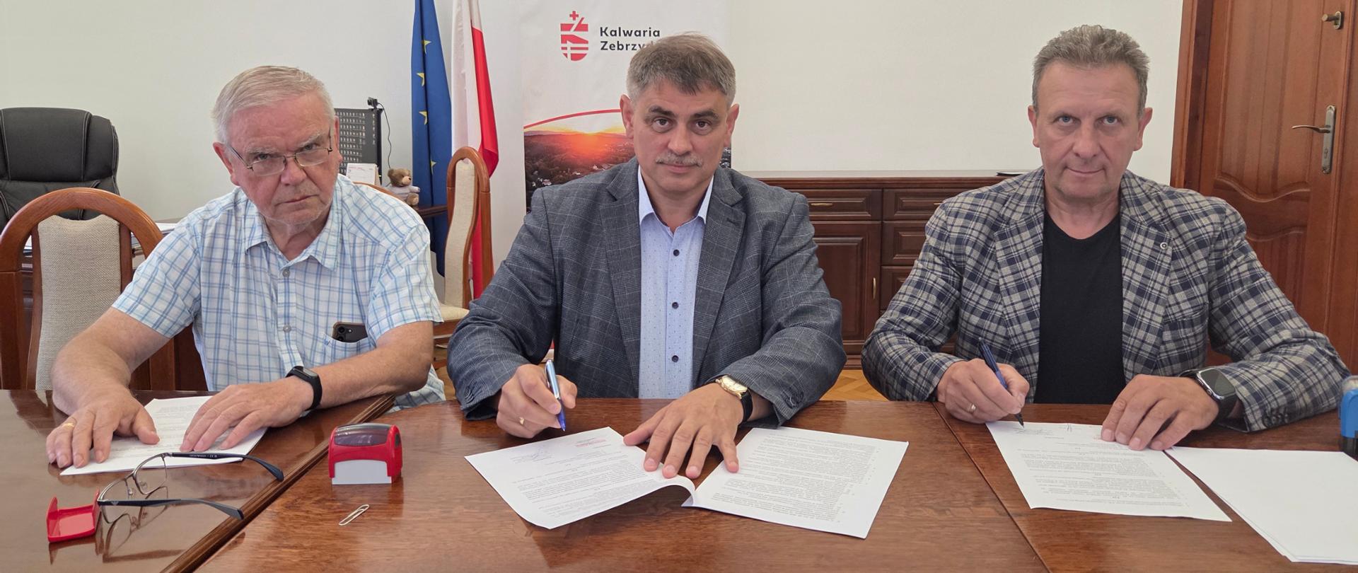 Na zdjęciu widać trzech mężczyzn siedzących przy drewnianym stole w sali Urzędu Miasta i Gminy Kalwaria Zebrzydowska podczas podpisywania dokumentów. W centralnej części kadru znajduje się Burmistrz Miasta Kalwarii Zebrzydowskiej, który podpisuje umowę, a po jego lewej i prawej stronie siedzą przedstawiciele wykonawcy oraz inspektora nadzoru, również składający podpisy na dokumentach. W tle widoczne są flagi Polski i Unii Europejskiej, roll-up z herbem i logo Gminy Kalwaria Zebrzydowska, godło Rzeczypospolitej Polskiej oraz obraz przedstawiający zabytkowy budynek. Zdjęcie dokumentuje oficjalne zawarcie umowy dotyczącej realizacji inwestycji.