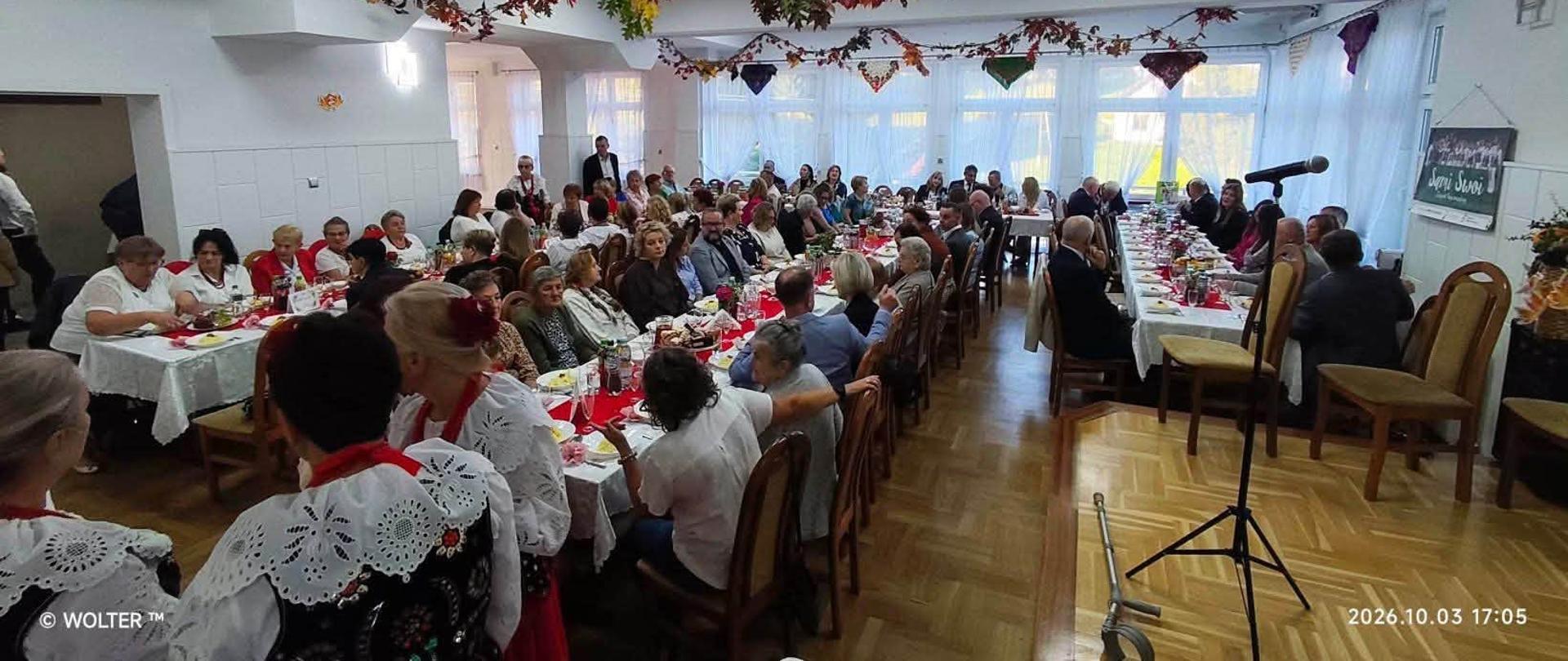Zdjęcie przedstawia uroczyste spotkanie jubileuszowe w sali wypełnionej gośćmi siedzącymi przy długich, odświętnie nakrytych stołach. W wydarzeniu uczestniczą m.in. członkowie zespołu w tradycyjnych strojach regionalnych, zaproszeni goście oraz przedstawiciele lokalnej społeczności. Sala udekorowana jest jesiennymi girlandami, a z prawej strony znajduje się mikrofon na statywie.