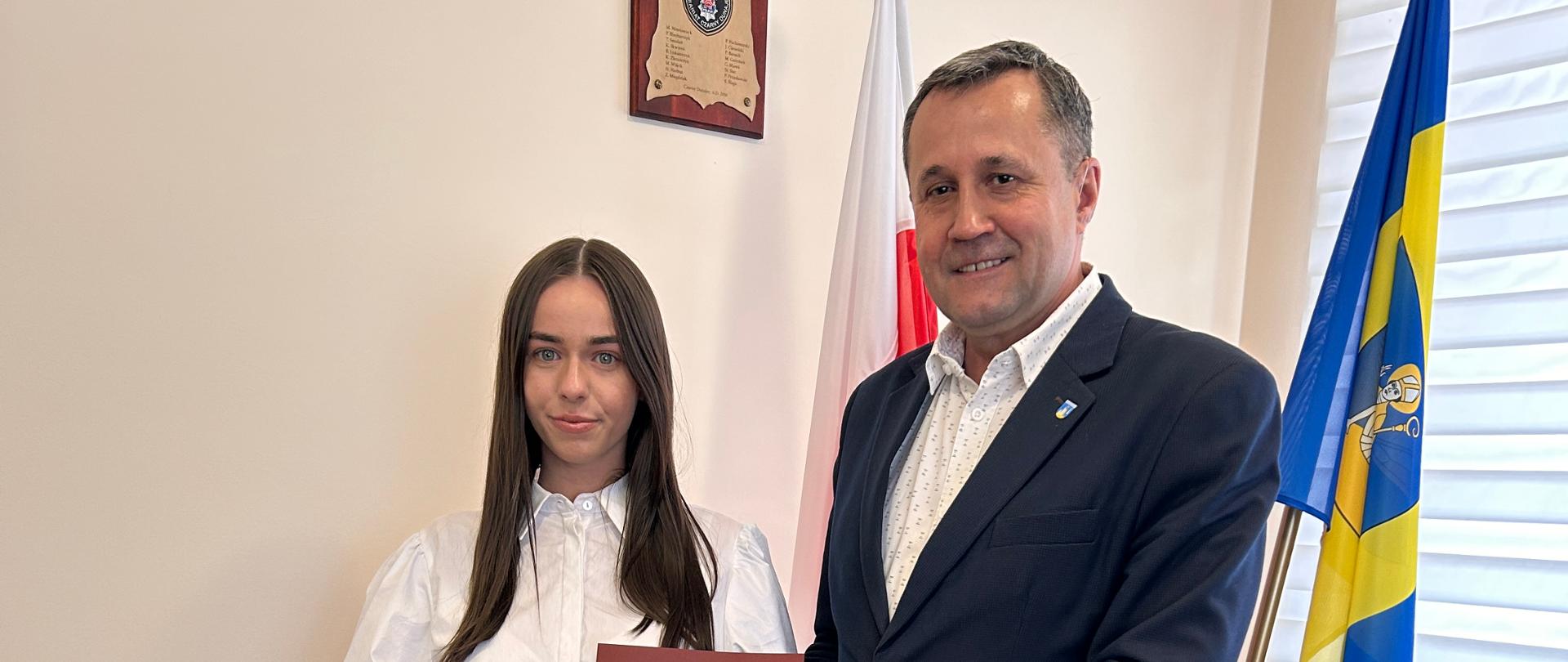 Mężczyzna i kobieta elegancko ubrani, pozują do zdjęć, trzymają w rękach bordową teczkę. W tle dwie flagi, kremowa ściana z obrazkiem.