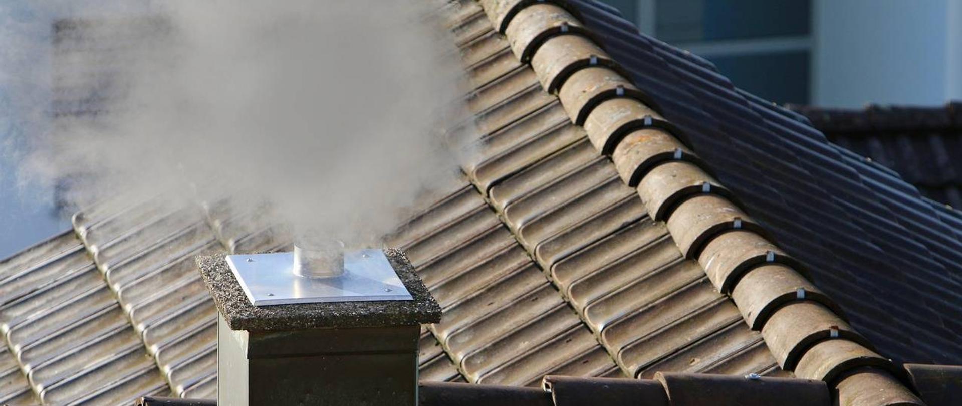 Dachówkowaty dach z kominem z którego wydobywa się ciemny dym