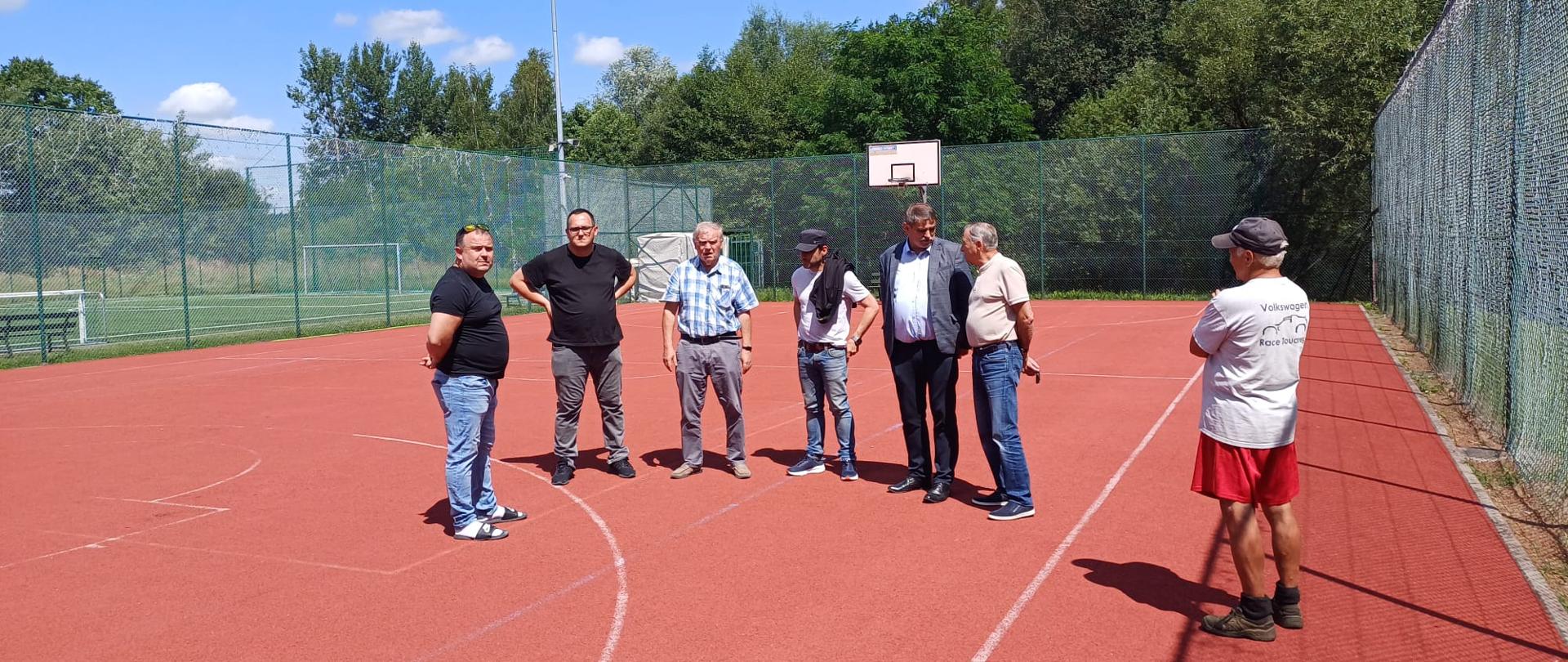 Siedmiu mężczyzn podczas oględzin wielofunkcyjnego boiska sportowego z czerwoną nawierzchnią. W tle widoczne są ogrodzenie, bramka, kosz do koszykówki, oświetlenie oraz zieleń.