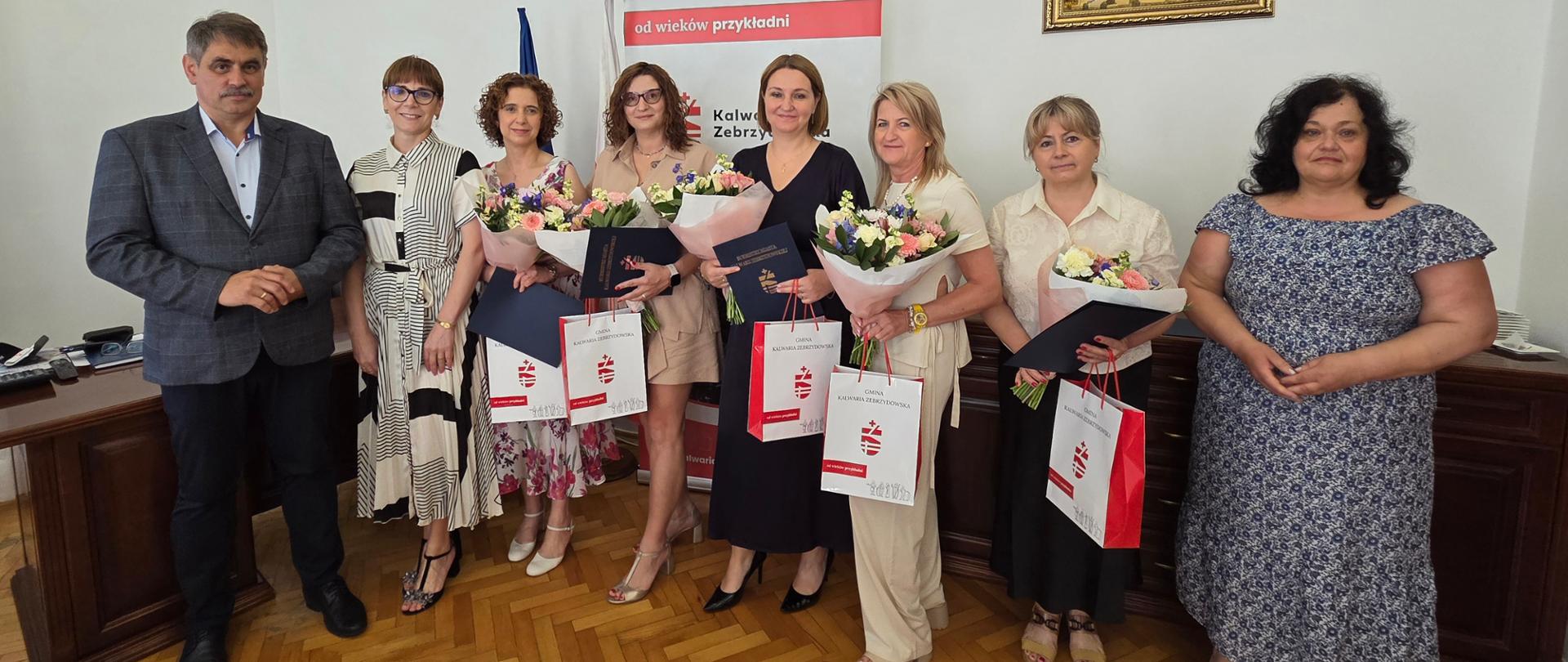 Burmistrz Miasta Kalwarii Zebrzydowskiej wraz z dyrektorami szkół podczas uroczystego wręczenia aktów powierzenia stanowisk na kolejne kadencje. Uczestnicy spotkania pozują do wspólnego zdjęcia, trzymając kwiaty, dokumenty oraz upominki.