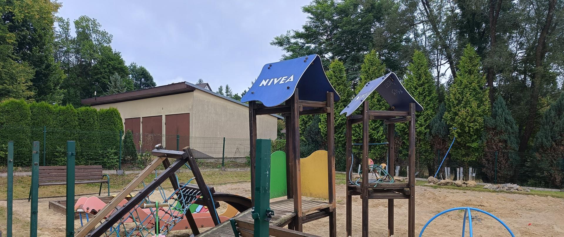 Plac budowy nowego placu zabaw w Skawie. zdemontowane urządzenia placu zabaw, W tle budynek, zielone drzewa, i jasno niebieskie niebo.