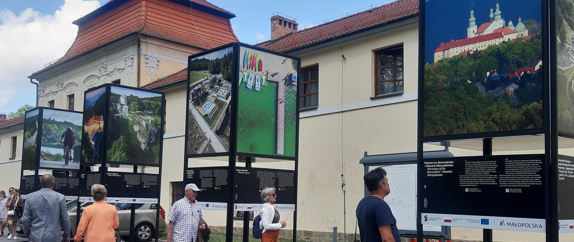 Zdjęcie przedstawia wystawę "Małopolska na Weekend z Funduszami Europejskimi. W tle wieże ze zdjęciami, wokół osoby. 