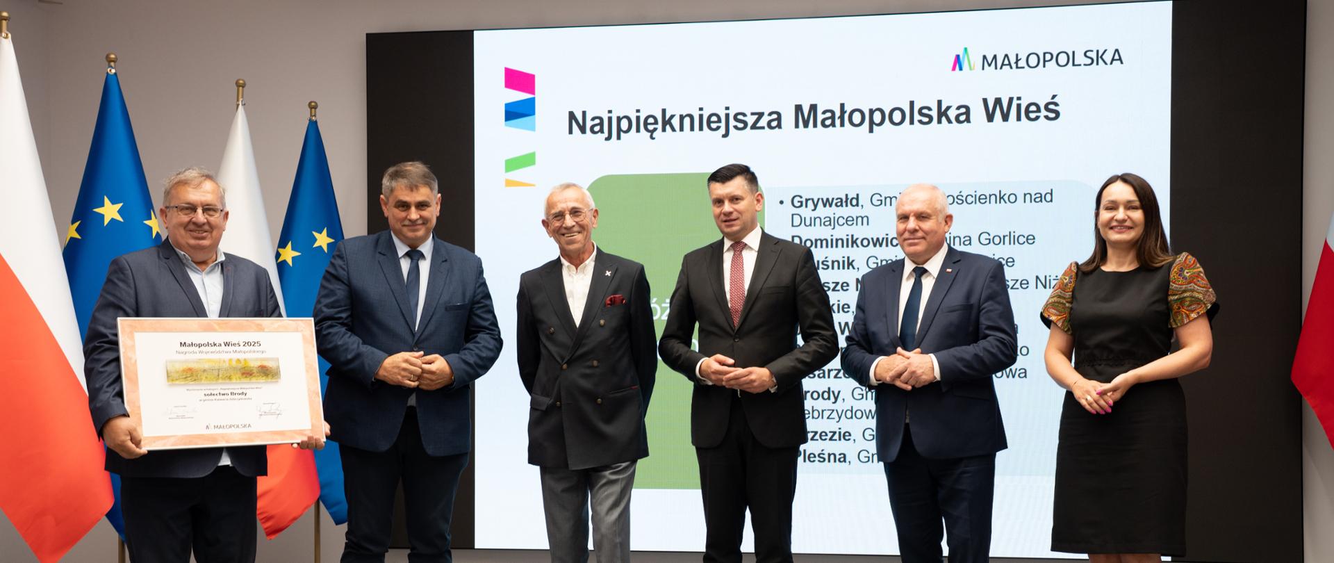 Uroczyste wręczenie tytułu 'Najpiękniejsza Małopolska Wieś 2025'. Na zdjęciu sześć osób stoi na tle ekranu z napisem 'Najpiękniejsza Małopolska Wieś'. Jedna z osób trzyma duży dyplom z nazwą zwycięskiej miejscowości. W tle widoczne flagi Polski i Unii Europejskiej oraz logo Małopolski