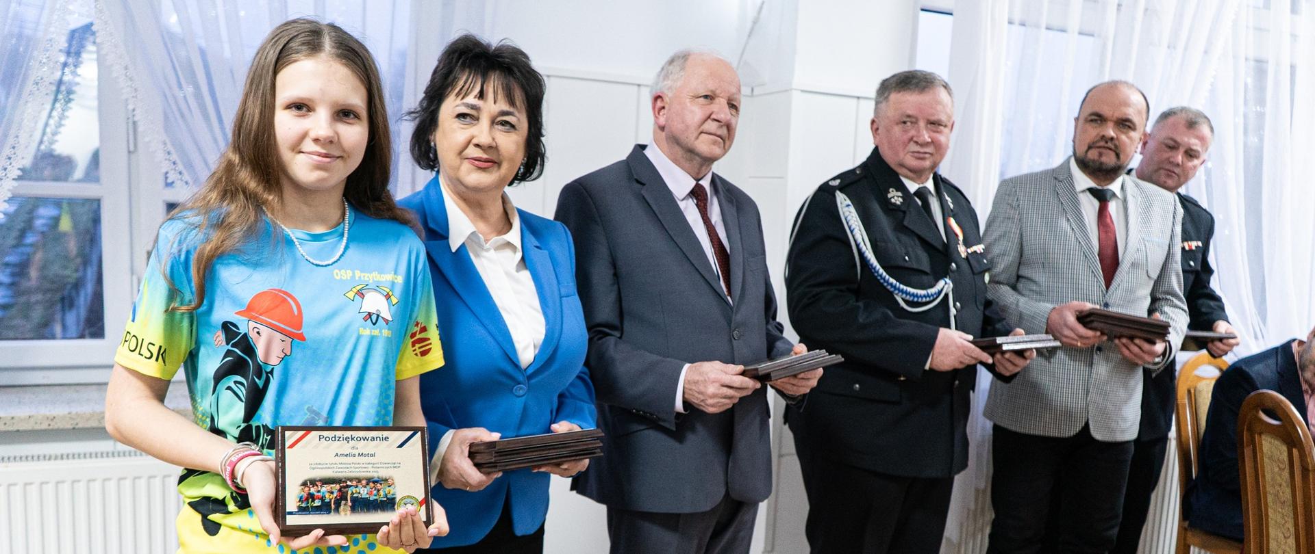 NA fotografii znajduje się sześć osób. Dziewczynka trzyma wyróżnienie za nią stoi kobieta w niebieskim kostiumie i białej bluzce, dalej mężczyzna w garniturze, kolejno strażak w odświętnym ubraniu mężczyzna w jasnej marynarce i czerwonym krawacie, na końcu strażak.