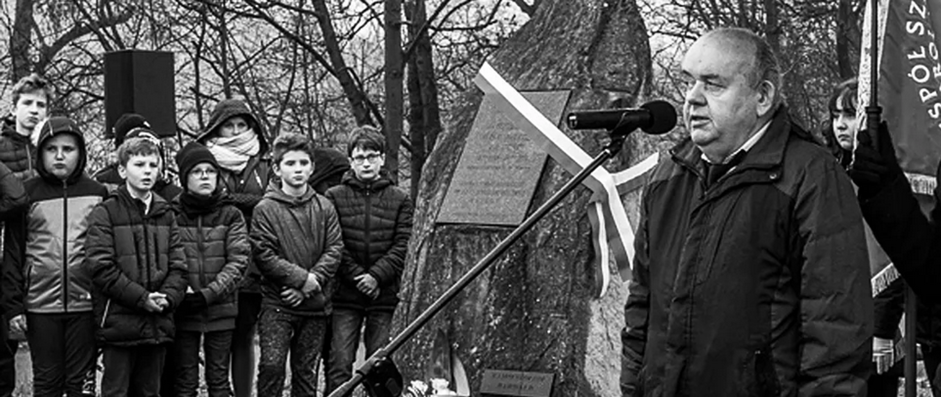 Czarno białe zdjęcie przedstawia dr. hab. Andrzeja Nowakowskiego przemawiającego do mikrofonu podczas uroczystości przy kamiennym obelisku z tablicą pamiątkową. W tle stoją uczniowie i opiekunowie ubrani w zimowe kurtki, tworząc skupioną grupę. Obok obelisku widoczna jest biało czerwona wstęga oraz zapalone znicze. Scena odbywa się w plenerze, wśród bezlistnych drzew.