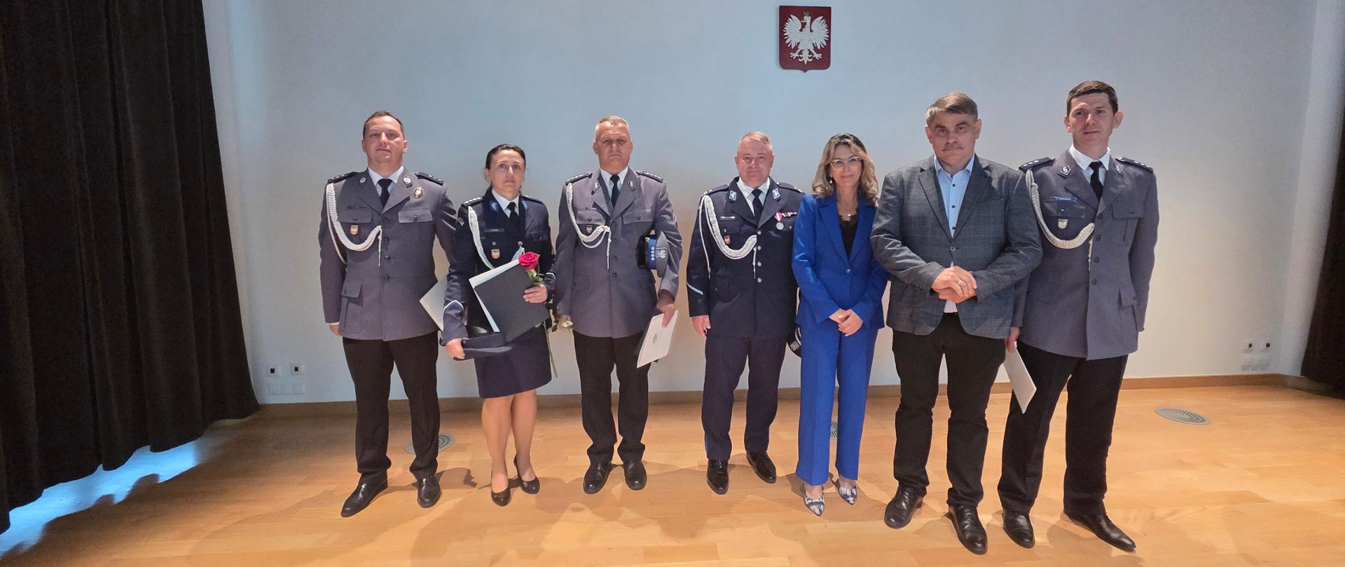 Siedem osób pozuje do wspólnego zdjęcia w sali pod godłem Polski. W grupie znajduje się pięcioro funkcjonariuszy Policji w mundurach oraz dwoje przedstawicieli samorządu. Część policjantów trzyma dyplomy, odznaczenia i różę.