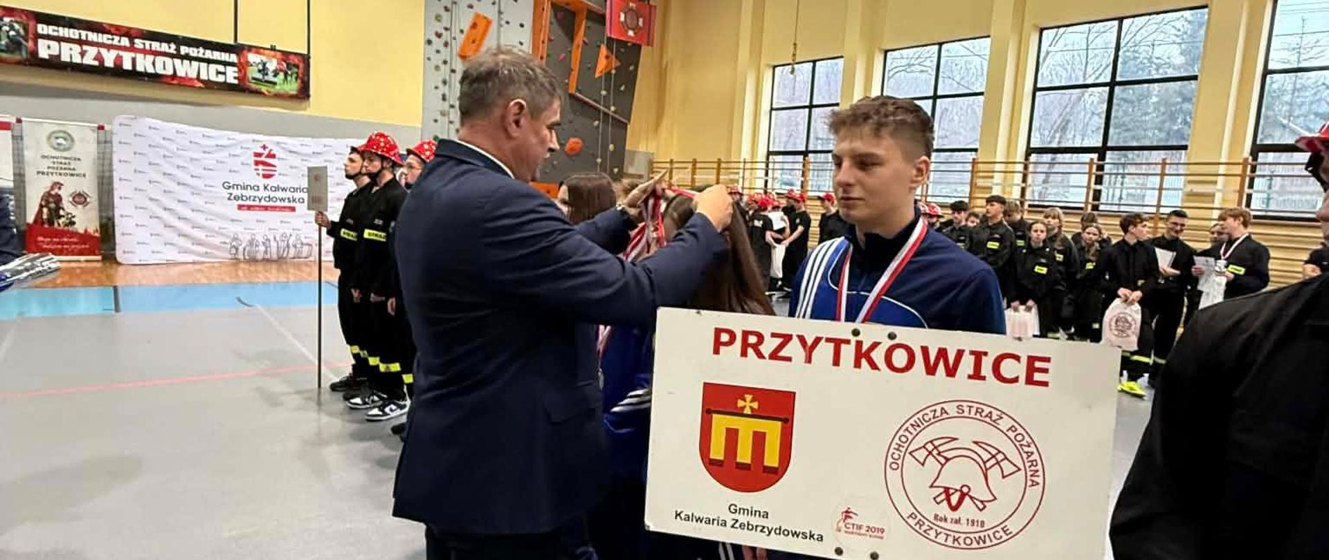 W hali sportowej podczas zawodów strażackich mężczyzna w garniturze zakłada medal młodemu zawodnikowi z drużyny OSP Przytkowice. Zawodnik trzyma tablicę z nazwą „Przytkowice” oraz herbem gminy i logotypem jednostki OSP. W tle widać pozostałych uczestników ustawionych w rzędach, ścianę wspinaczkową oraz banery z informacją o wydarzeniu. Atmosfera uroczysta, związana z wręczaniem nagród.