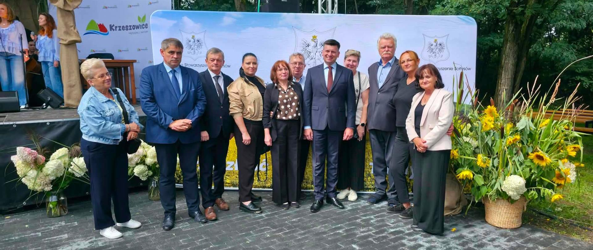Grupowe zdjęcie uczestników Małopolskiego Spotkania Sołtysów, wykonane przed plenerową sceną w Zalasie w Gminie Krzeszowice. W tle widoczna jest ścianka wydarzenia, a po bokach kompozycje z hortensji i słoneczników.