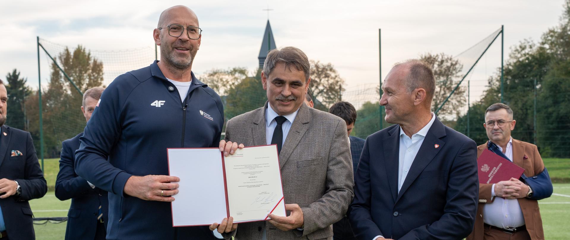 Na boisku sportowym w Kalwarii Zebrzydowskiej trzech mężczyzn pozuje do zdjęcia podczas uroczystości związanej z przyznaniem dofinansowania na modernizację kompleksu „Orlik”. Jeden z nich trzyma otwartą teczkę z dokumentem potwierdzającym przyznanie środków. W tle widoczni są pozostali uczestnicy wydarzenia oraz infrastruktura boiska sportowego.