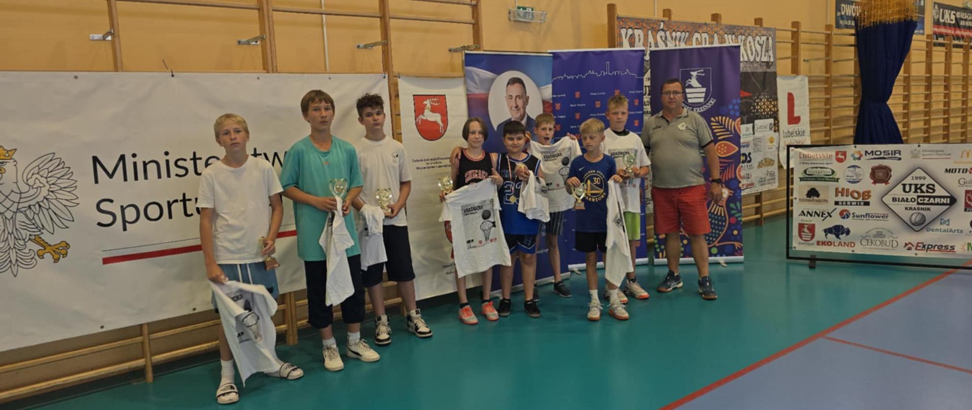 Ośmiu młodych zawodników wyróżnionych w jednej z kategorii wiekowych, trzymający w rękach otrzymane koszulki i statuetki. Zawodnicy stoją na hali sportowej przy ścianie, na tle banerów partnerów turnieju. Pierwszy od prawej stoi prezes Uczniowskiego Klubu Sportowego Biało-Czarni w Kraśniku.
